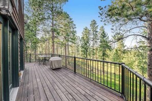 Eine Holzterrasse mit Tisch und Stühlen, umgeben von einem schwarzen Geländer, mit Blick auf ein bewaldetes Gebiet.
