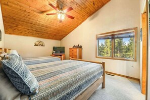 Ein Schlafzimmer mit Holzbalkendecke, einem Bett mit gestreifter Bettdecke, einem Fernseher, einer Kommode und einem Fenster mit Blick auf Bäume.
