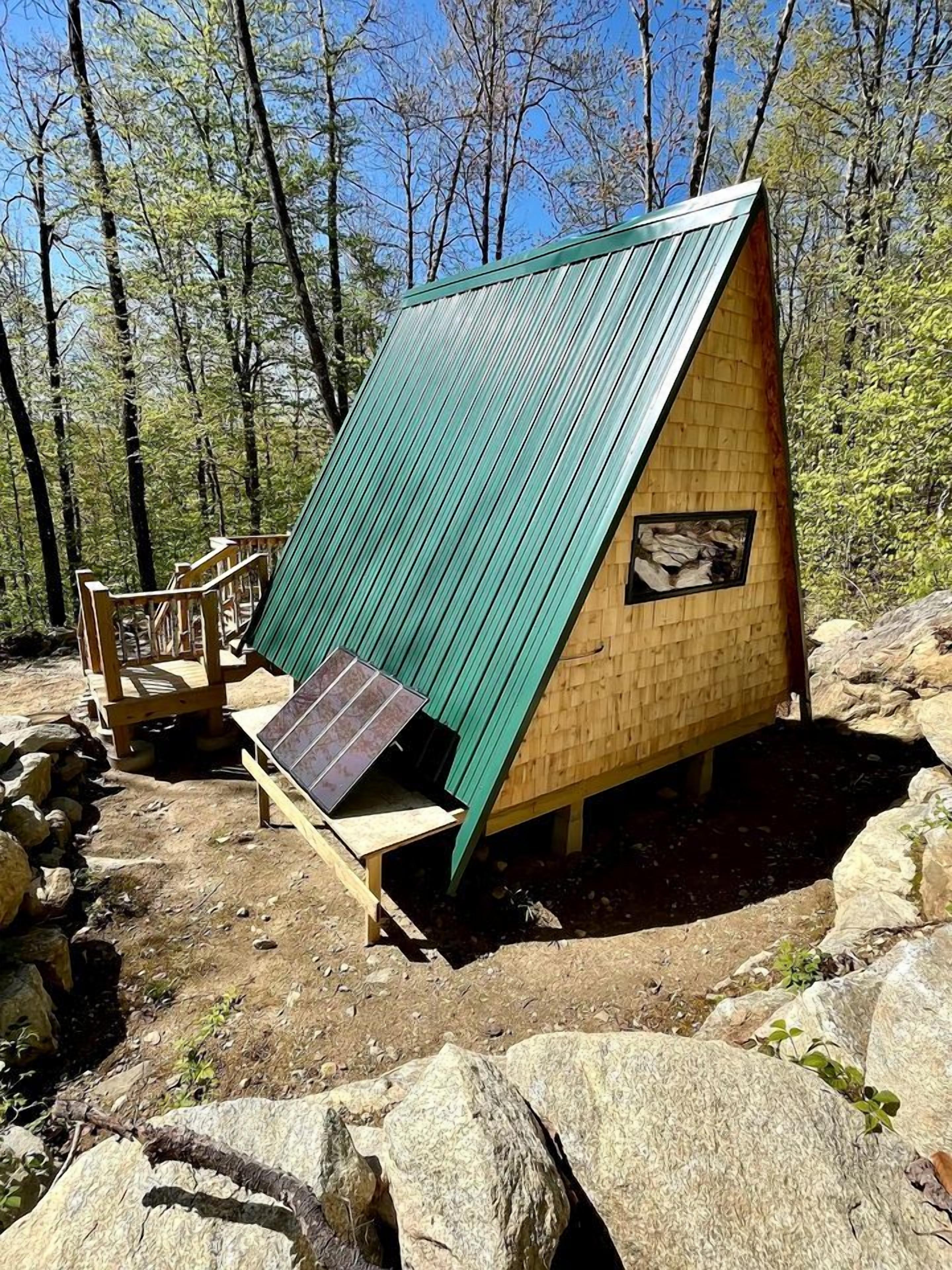 A small wooden cabin with a green roof, a solar panel, and a wooden deck.