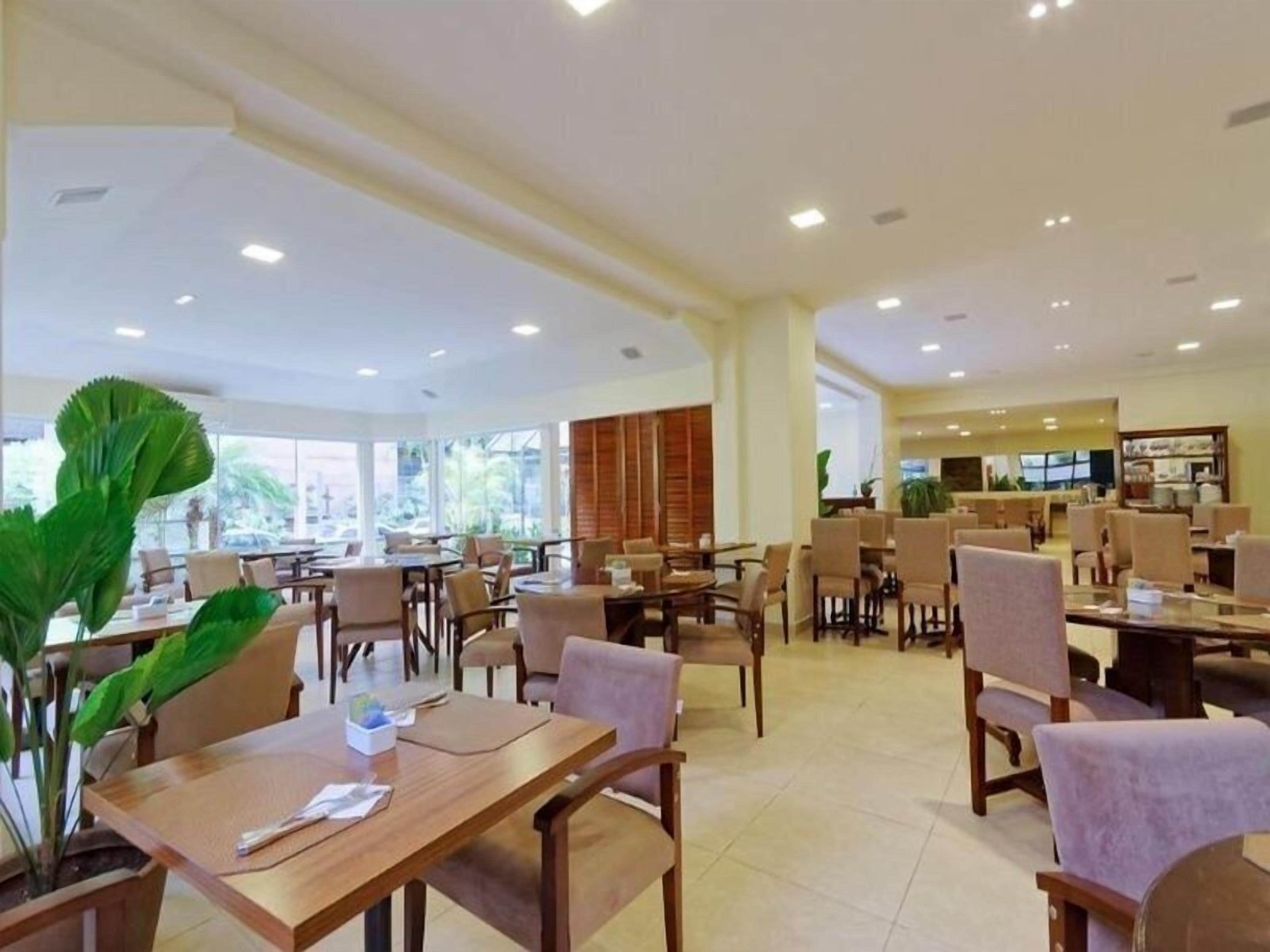 Un vaste espace dédié à la salle à manger, avec de nombreuses tables et chaises, de grandes fenêtres et des plantes d’intérieur.