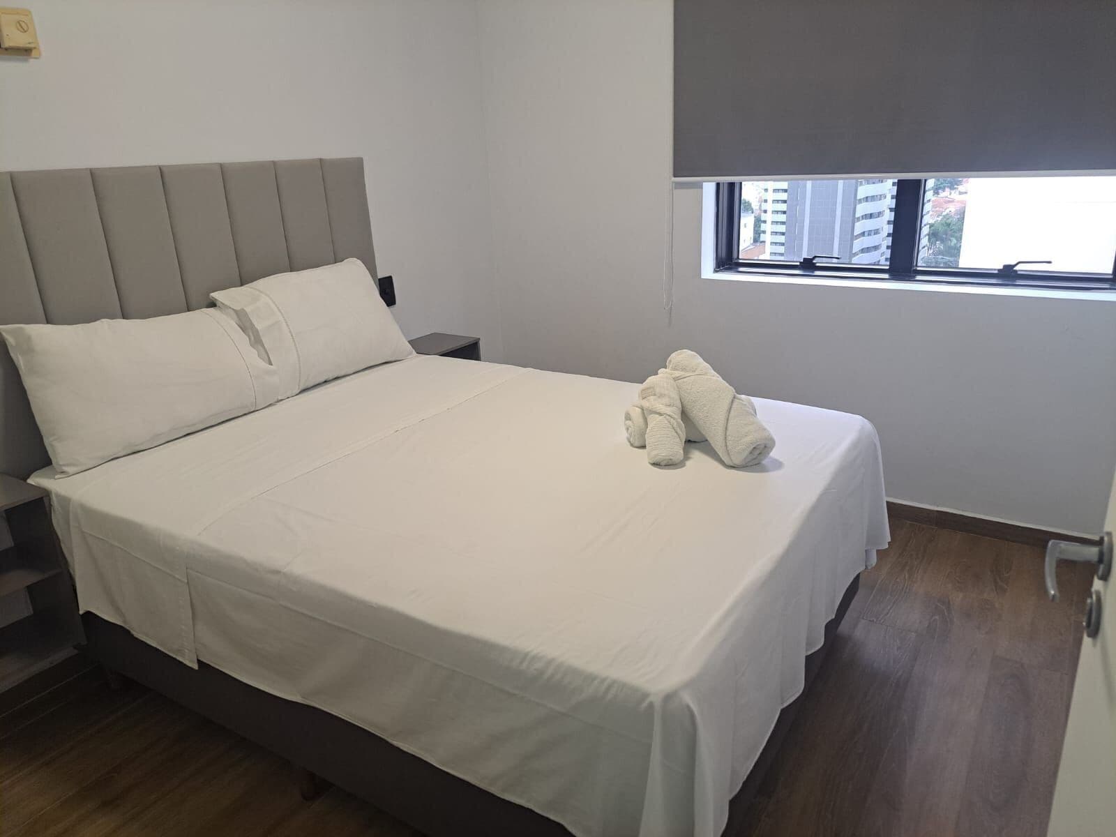 Quarto de hotel com cama, mesa de cabeceira, janela com vista para edifícios e uma persiana pequena.