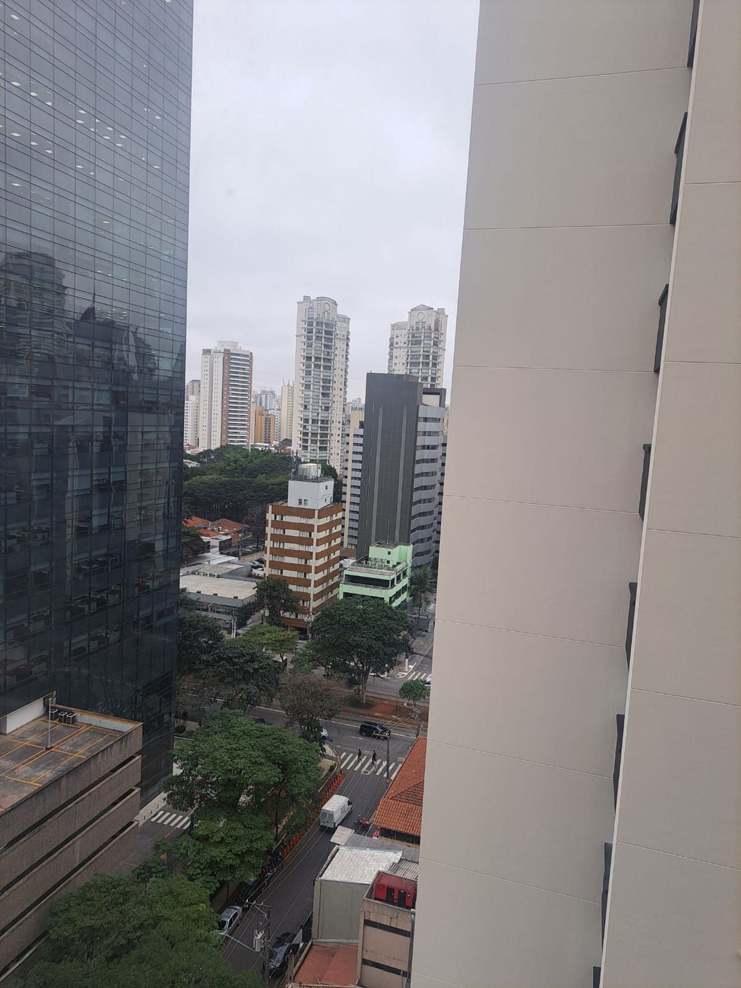 Um edifício alto com fachada de vidro moderna, rodeado por outros edifícios altos e uma rua com veículos.