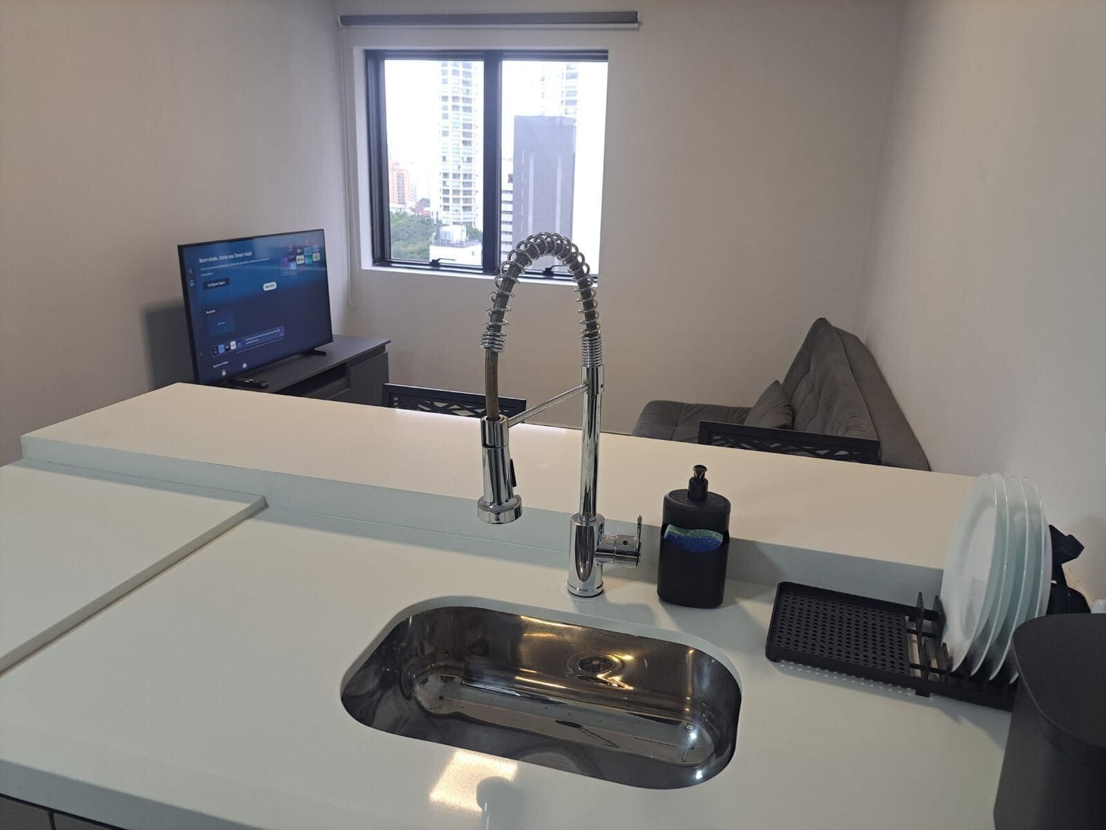 A modern kitchen with a stainless steel sink, a faucet, a window with a view of buildings, and a computer on a desk.