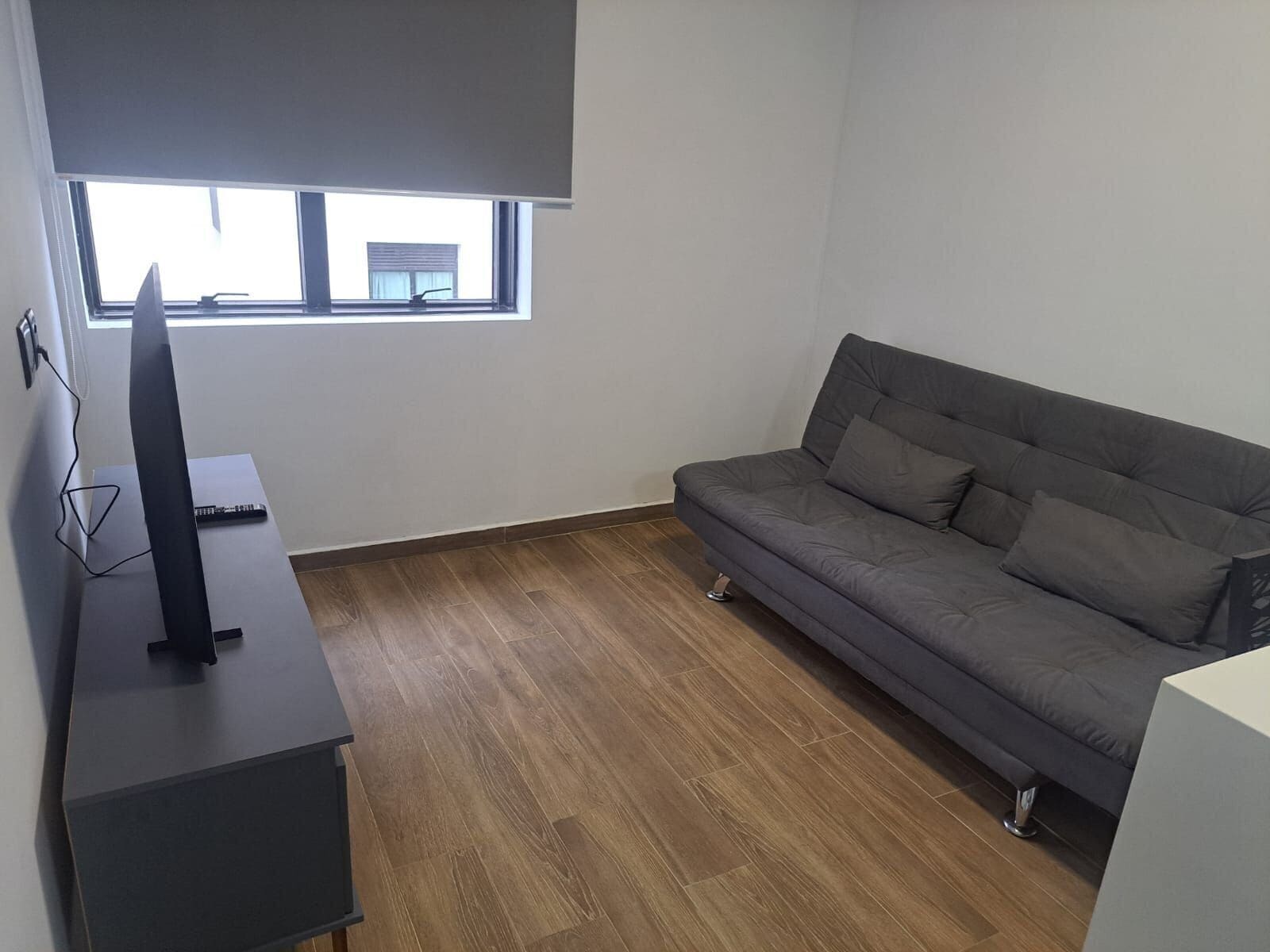 A room with a grey sofa, a flat-screen TV on a stand, and a window with blinds.