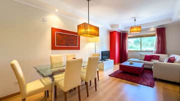 A modern living room with a glass dining table, white sofas, and a red accent wall.