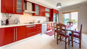 A modern kitchen with red cabinets, a dining area with wooden chairs, and a view through the window. - Ref gio do Mar by Acasadascasas (Mafra)