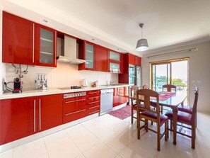 A modern kitchen with red cabinets, a dining area with wooden chairs, and a view through the window. - Ref gio do Mar by Acasadascasas (Mafra)