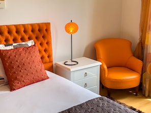 Ein Hotelzimmer mit einer gepolsterten Kopfstütze, einem Nachttisch mit Lampe, einem Sessel und einem Bett mit gemustertem Kopfkissen.