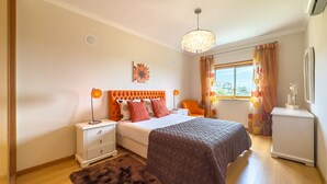 A bedroom with a bed, nightstands, a dresser, a chandelier, and a window with curtains. - Ref gio do Mar by Acasadascasas (Mafra)