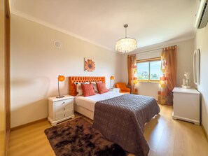 A bedroom with a bed, nightstands, a dresser, a chandelier, and a window with curtains. - Ref gio do Mar by Acasadascasas (Mafra)