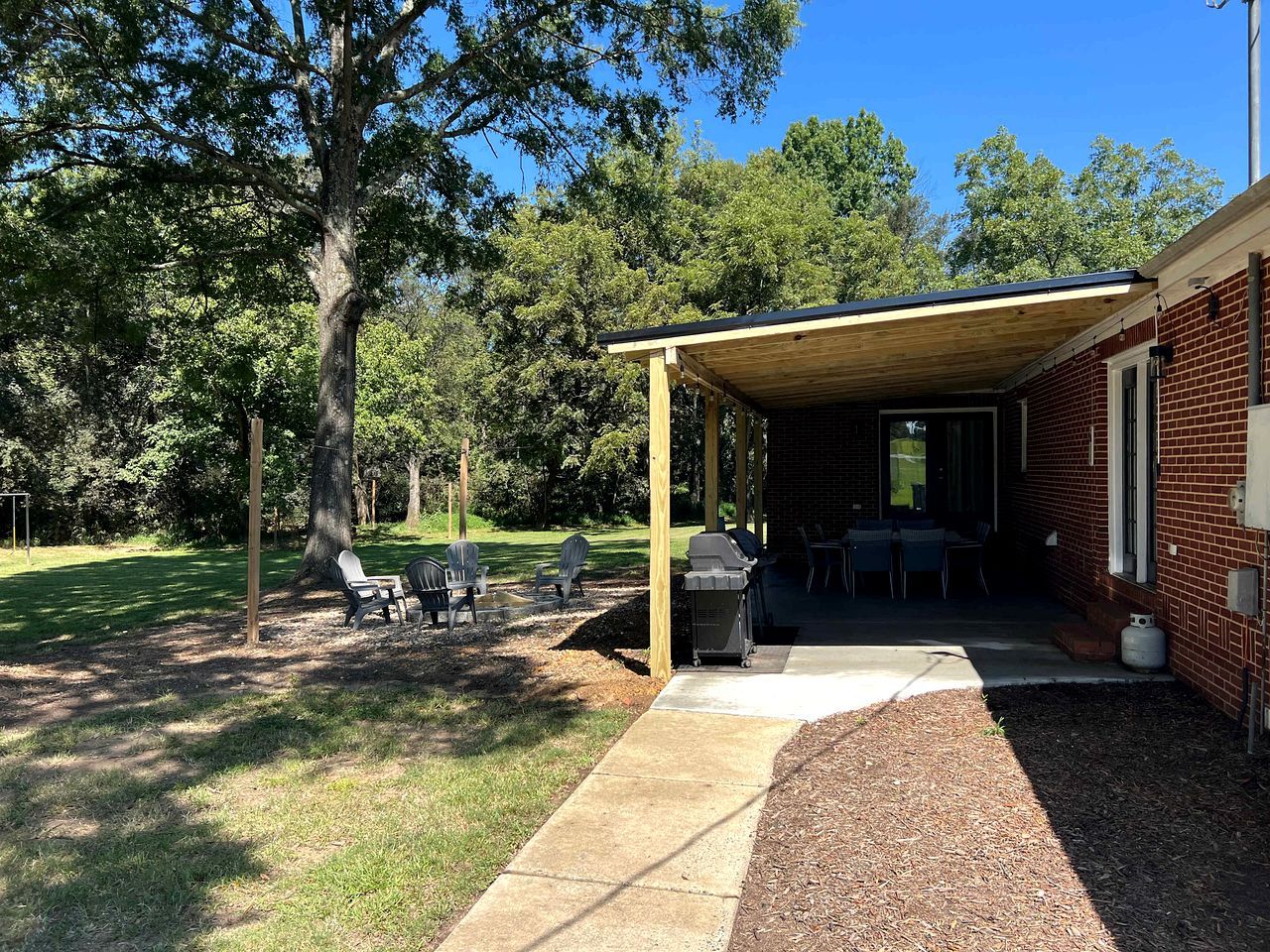A covered outdoor area with a barbecue grill, chairs, and a concrete pathway leading to a building.