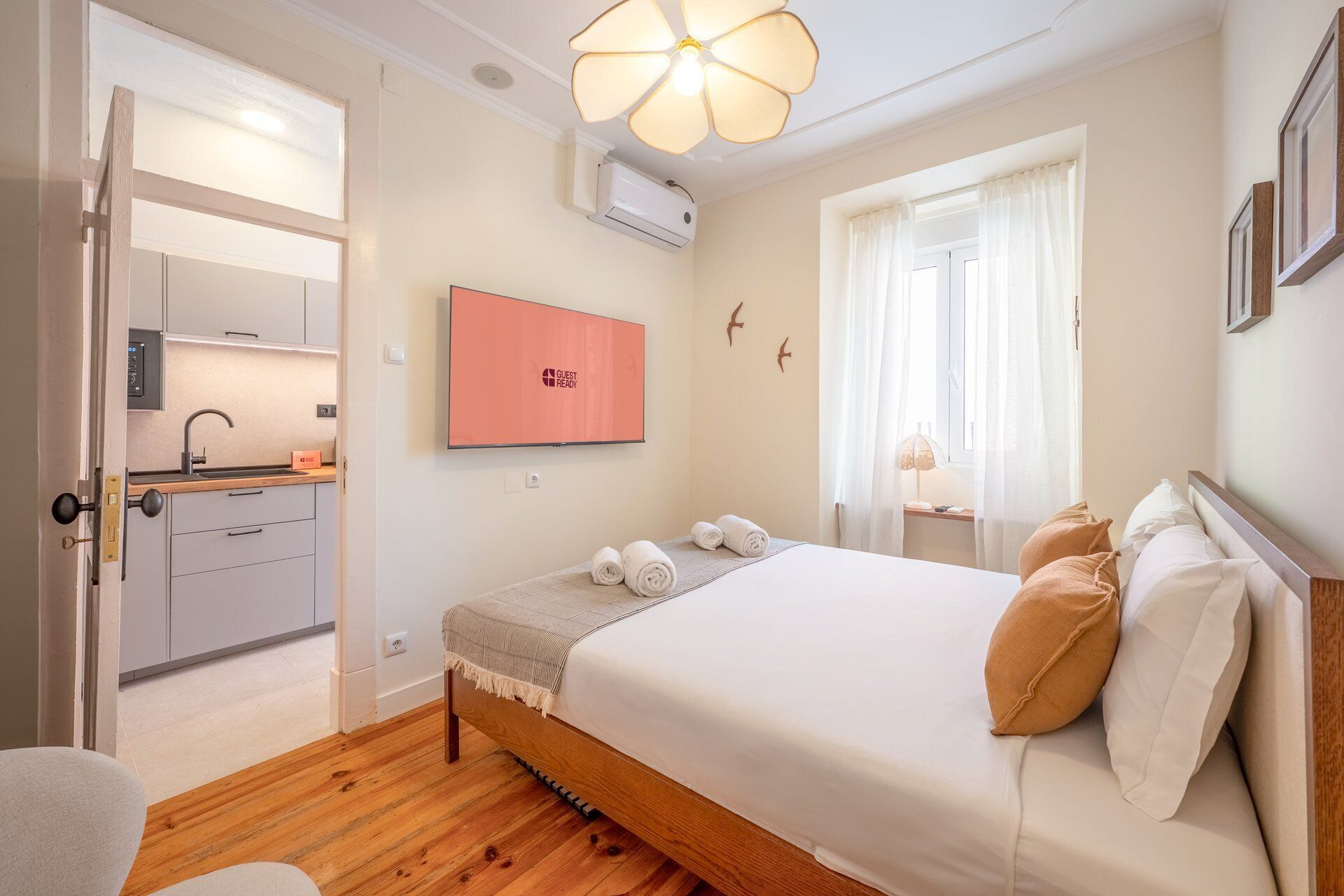 A hotel room with a bed, a desk, a chair, a window with curtains, a wall-mounted TV, and a door leading to a kitchenette.