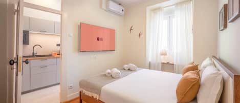 A hotel room with a bed, a desk, a chair, a window with curtains, a wall-mounted TV, and a door leading to a kitchenette.