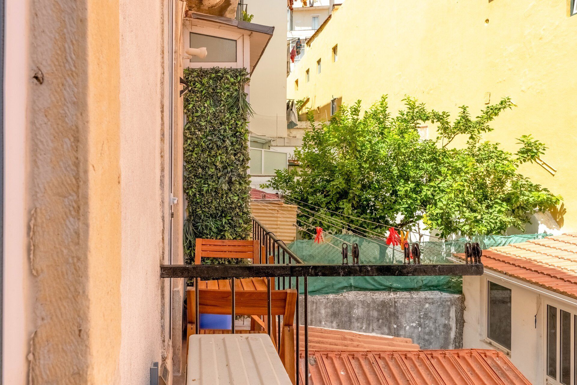 A narrow urban street with a wooden bench, a metal railing, and a tree providing shade.