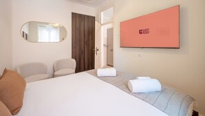 Una habitación de hotel con cama, dos sillas, un espejo, televisor montado en la pared y una puerta que da a otra estancia.