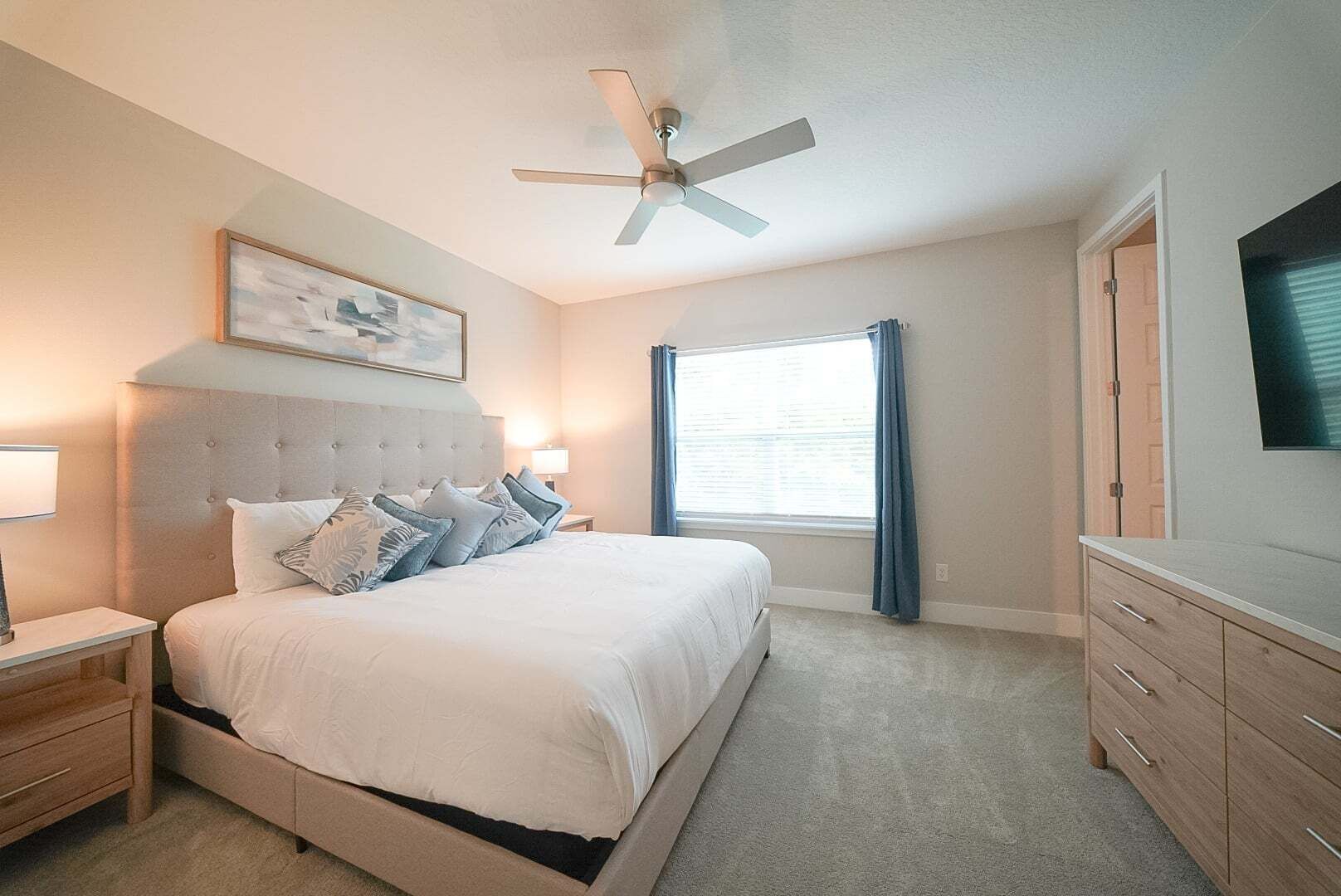 Une chambre à coucher comprenant un lit, des tables de chevet, un ventilateur de plafond, une fenêtre avec des rideaux et un téléviseur posé sur une commode.