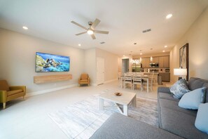 A modern living room with a flat-screen TV, a ceiling fan, and a dining area with a kitchen in the background. - Royal View Retreat-1212 (Kissimmee)