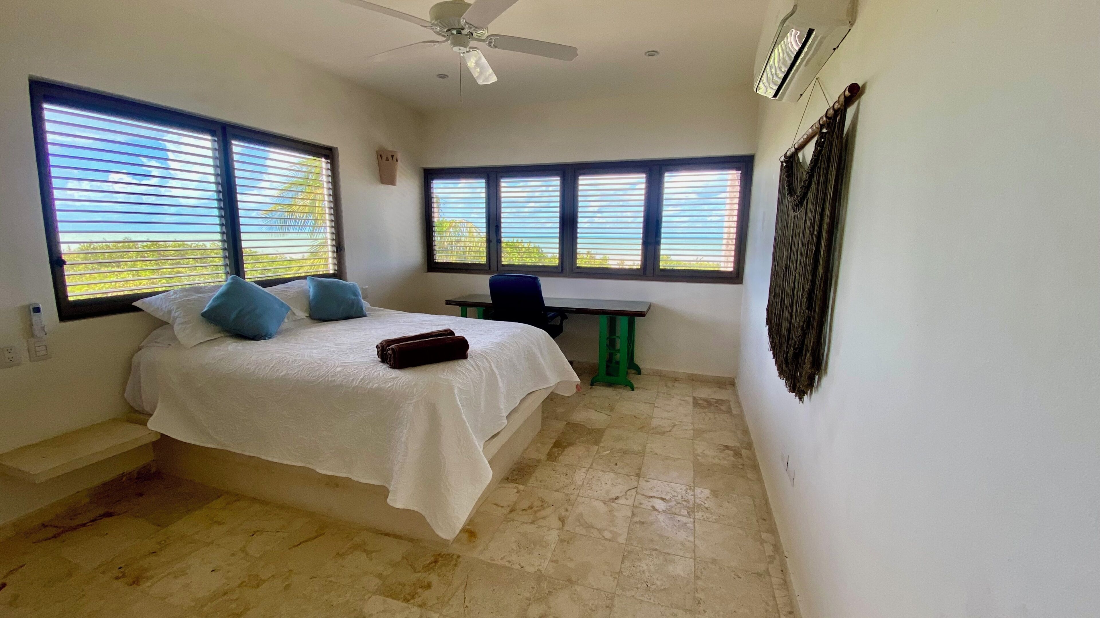 A bedroom with a bed, a desk, and a chair. There are windows with blinds, a ceiling fan, and an air conditioning unit.