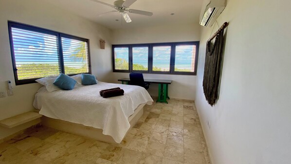 A bedroom with a bed, a desk, and a chair. There are windows with blinds, a ceiling fan, and an air conditioning unit.