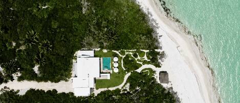 An aerial view of a property with a swimming pool, surrounded by dense greenery and a sandy beach.