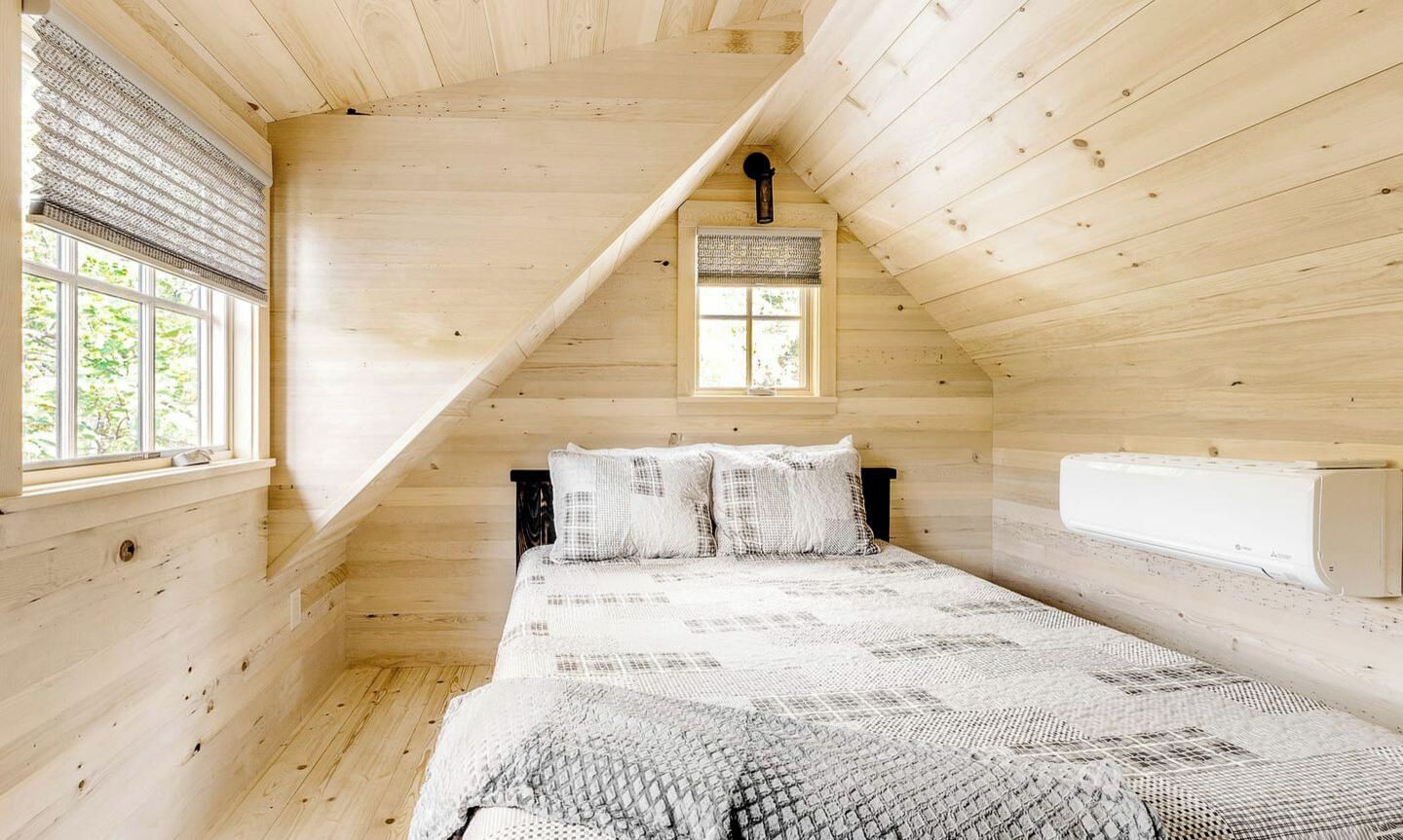 A room with a sloped ceiling, a bed with a patterned bedspread, a window with blinds, and a wall-mounted heater.