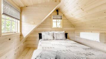A room with a sloped ceiling, a bed with a patterned bedspread, a window with blinds, and a wall-mounted heater.