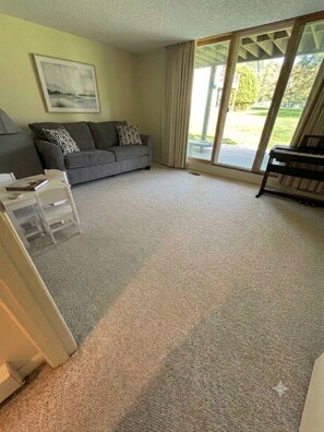 A living room with a grey sofa, a piano, and a large sliding glass door leading to a balcony. - Family friendly condo at Smugglers Notch Resort (Cambridge)