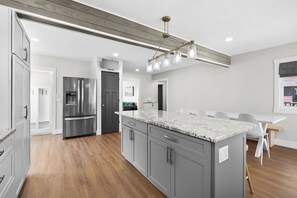 A modern kitchen with grey cabinetry, a stainless steel refrigerator, a marble countertop, and a dining area with white chairs. - Paddle & Play on Long Lake - Lakefront Retreat (Scotts)