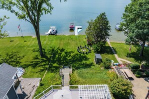 A lakeside property with a grassy lawn, a dock with boats, and a small deck with outdoor seating. - Paddle & Play on Long Lake - Lakefront Retreat (Scotts)