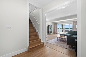A modern interior with a wooden staircase, a living area with a sofa and a coffee table, and a view through large windows. - Paddle & Play on Long Lake - Lakefront Retreat (Scotts)