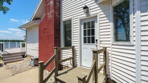 A house with a red brick chimney, white siding, a front door, and a wooden deck with steps leading up to it. - Paddle & Play on Long Lake - Lakefront Retreat (Scotts)