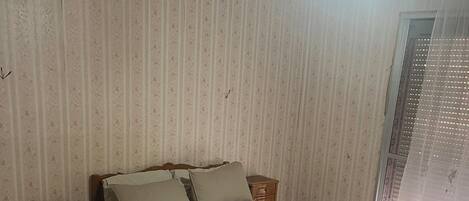 A bedroom with a bed, a nightstand, a patterned floor, and a window with a curtain.