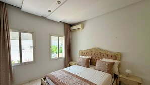Quarto com cama grande, duas mesas de cabeceira, ar-condicionado e janela com cortinas.
