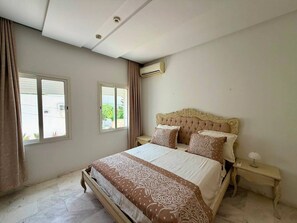 Ein Schlafzimmer mit einem großen Bett, zwei Nachttischen, einer Klimaanlage und einem Fenster mit Vorhängen.