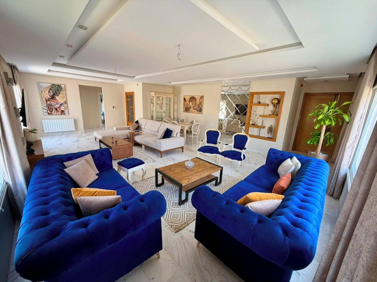 A modern living room with two large blue sofas, a coffee table, and a dining area with chairs.