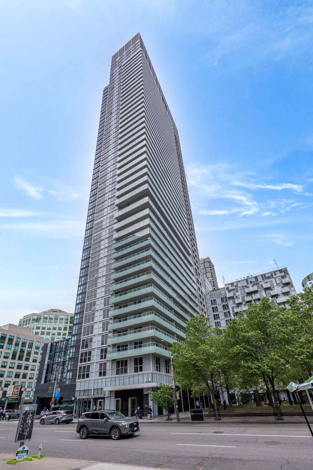A tall, modern skyscraper with a grid-like facade, surrounded by urban buildings and a street with cars and pedestrians.