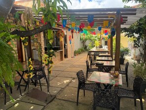 Un patio al aire libre con mesas y sillas, rodeado de plantas en macetas y un muro de ladrillo.