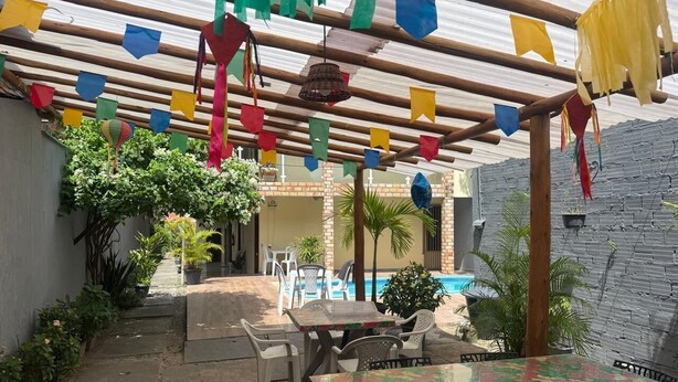 Un patio al aire libre con una mesa y sillas, rodeado de plantas en macetas y una piscina al fondo.