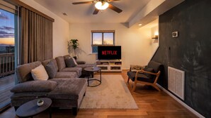Een moderne woonkamer met een hoekbank, een klein rond tafeltje, een houten vloer, een plafondventilator en een televisie waarop 'NETFLIX' staat.
