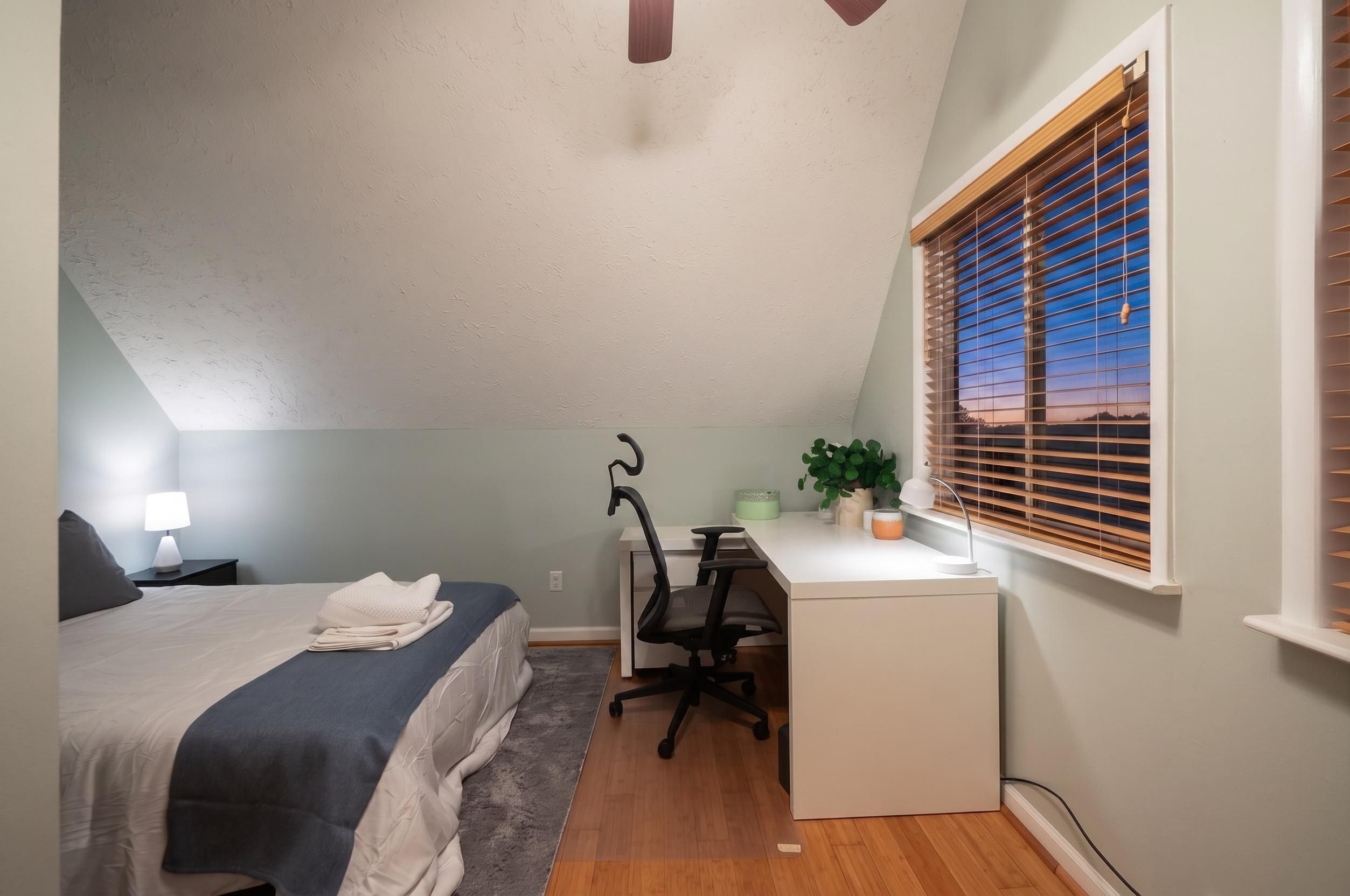 A bedroom with a bed, desk, chair, and a window with blinds.