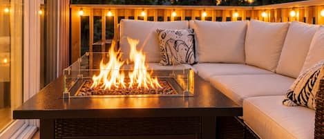 Una fogata con cerramiento de vidrio, un sofá de mimbre con cojines, y una terraza de madera con luces colgantes.