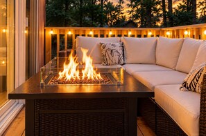A fire feature with a glass enclosure, a wicker sofa with cushions, and a wooden deck with string lights.