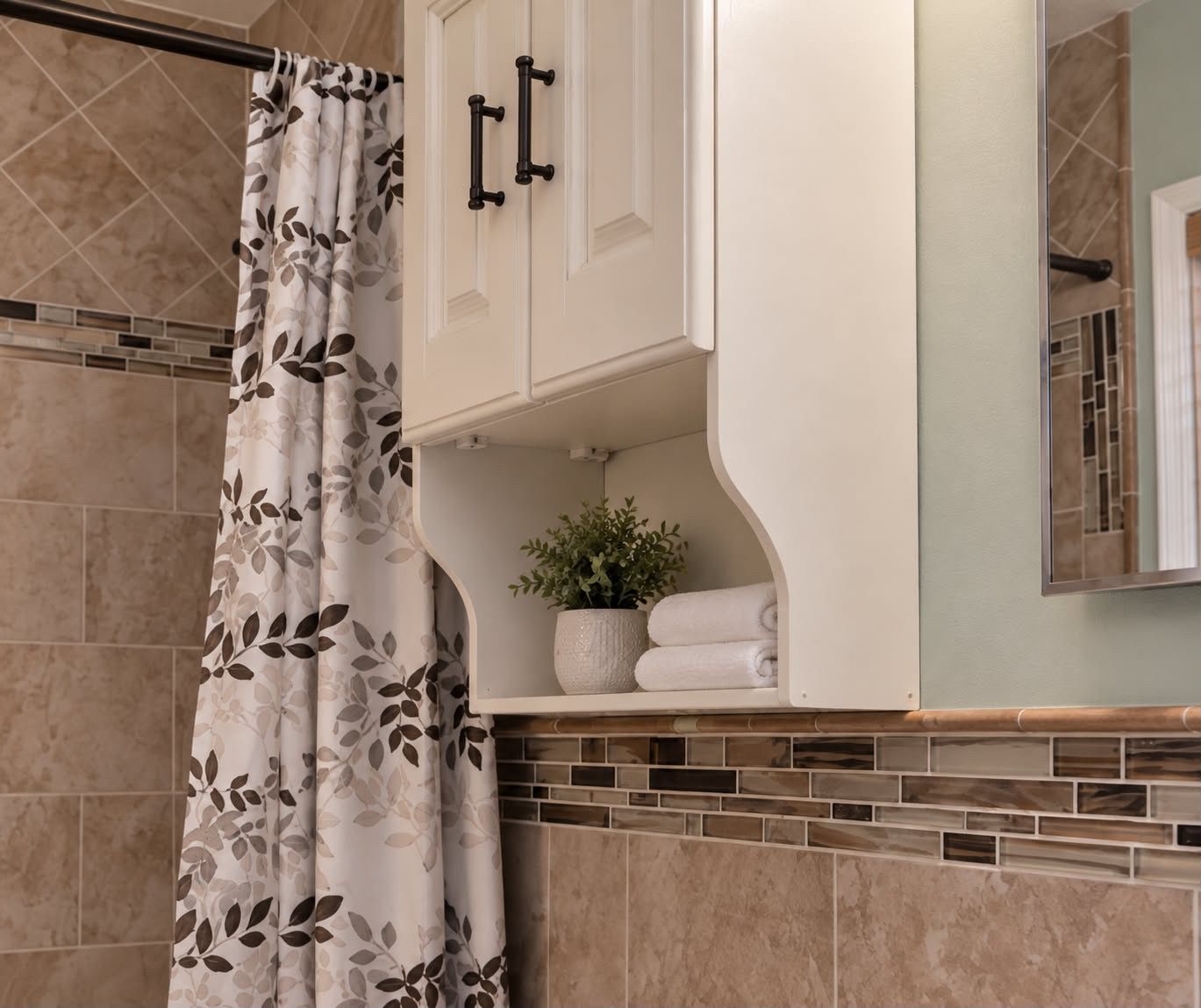 A bathroom with a shower curtain, a white cabinet with black handles, a potted plant, and a towel rack.