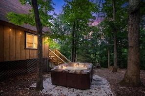 Uma banheira de hidromassagem em área arborizada, com uma casa de madeira e uma escadaria que leva a um deck.