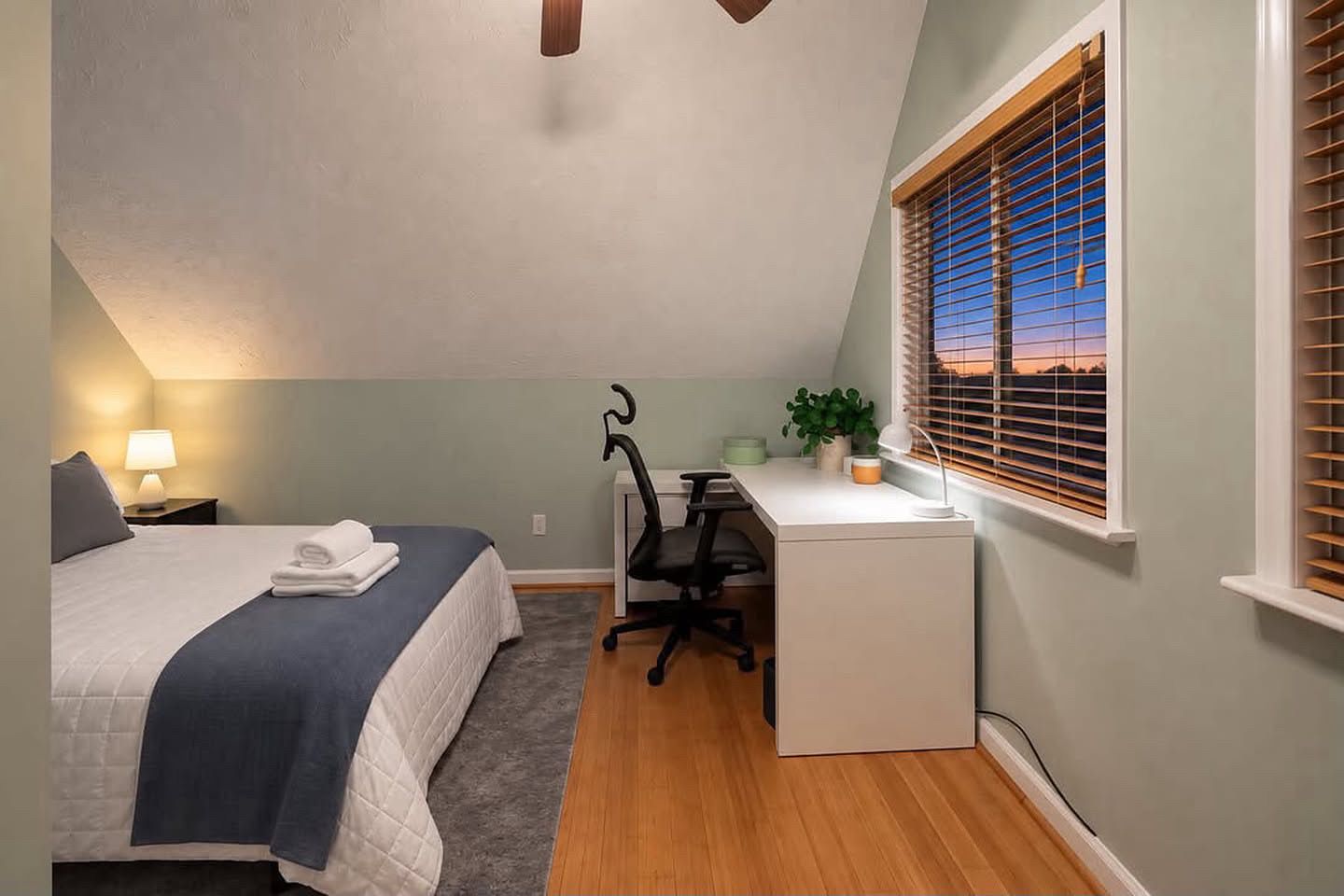 A bedroom with a bed, desk, chair, and a window with blinds.