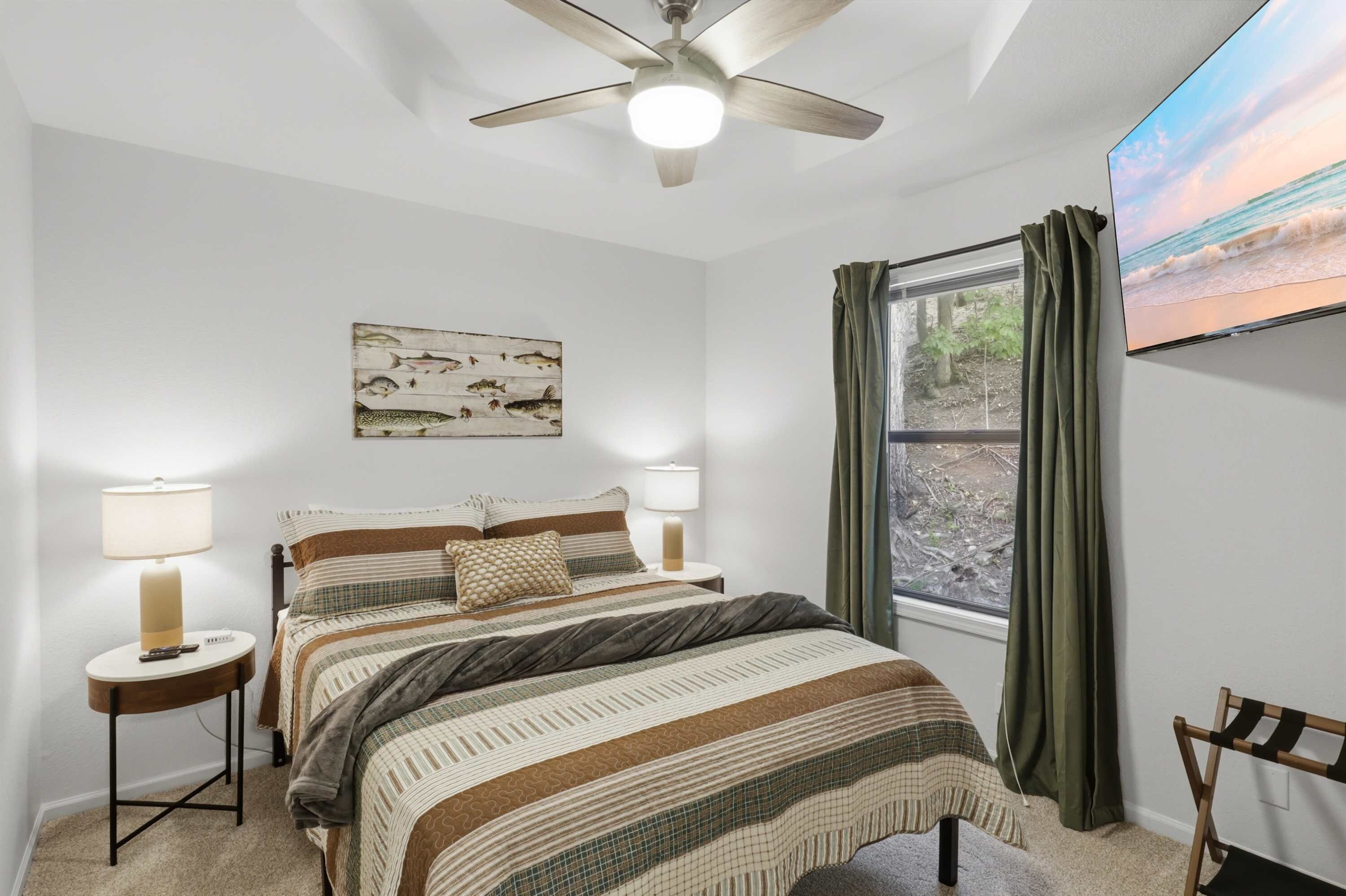 Une chambre à coucher avec un lit, deux tables de chevet, un ventilateur de plafond, un tableau, une fenêtre avec des rideaux et un téléviseur fixé au mur.
