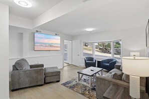 Una sala de estar moderna con un televisor de pantalla plana, sofás grises, una mesa de centro y vistas al exterior a través de amplios ventanales.