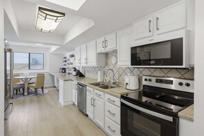 A modern kitchen with white cabinetry, a built-in microwave, a dishwasher, and an oven. There is a dining area with a table and chairs visible through an open door. - Lakefront 7BR retreat: Hot Tub Game Room Fire Pit (Lake Ozark)