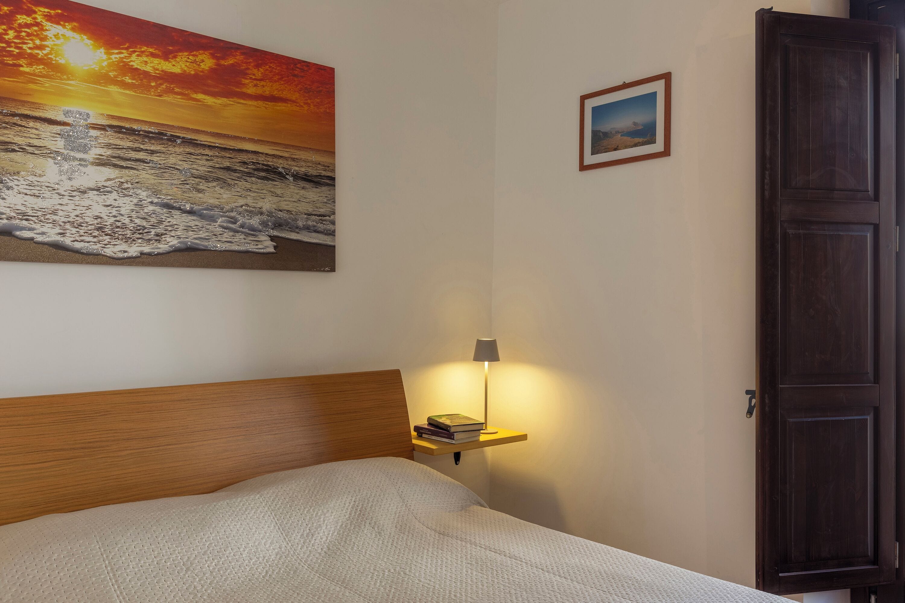 A bedroom with a bed, a nightstand with a lamp, a framed picture on the wall, and a door.
