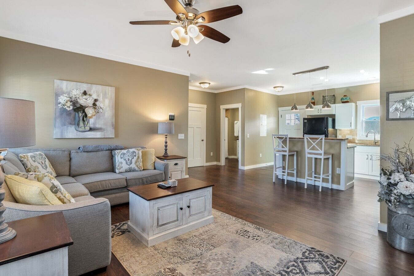 A spacious living room with a sofa, coffee table, and a ceiling fan. There is a kitchen area with a bar and stools.
